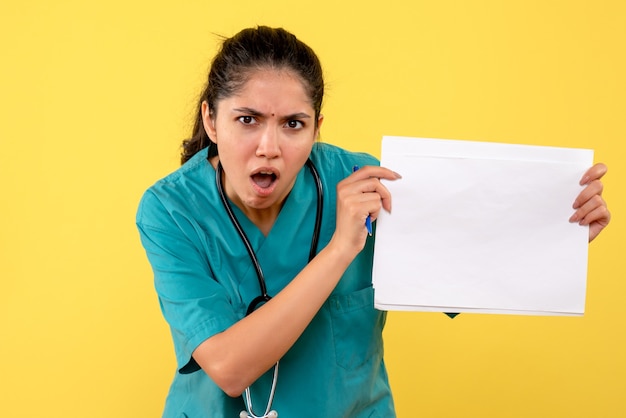 Vue de face de femme médecin avec bouche ouverte montrant des papiers sur mur jaune