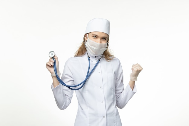 Vue de face femme médecin en combinaison médicale portant un masque en raison de coronavirus tenant un stéthoscope sur un mur blanc virus pandémique covid- maladie