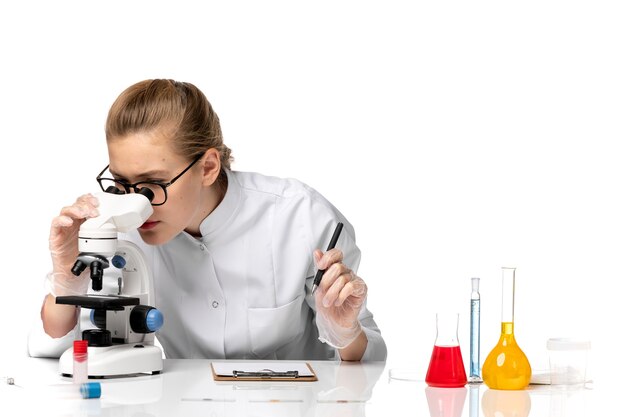 Vue de face femme médecin en costume médical blanc à l'aide d'un microscope sur un espace blanc