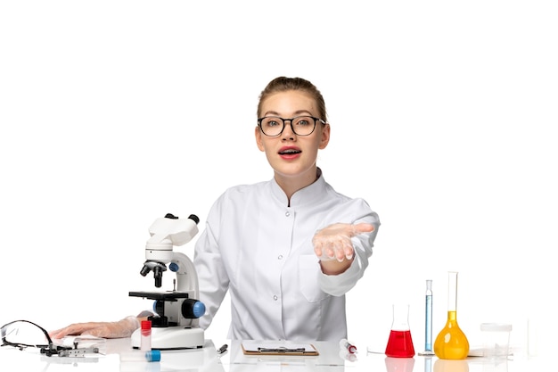 Vue de face femme médecin en costume médical blanc assis en face de la table avec des solutions sur un bureau blanc