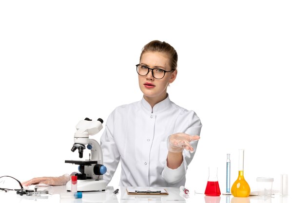 Vue de face femme médecin en costume médical blanc assis en face de la table avec des solutions sur l'espace blanc