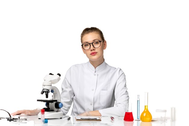 Vue de face femme médecin en costume médical blanc assis en face de la table avec des solutions sur l'espace blanc