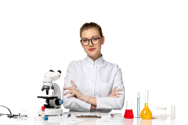 Vue de face femme médecin en costume médical blanc assis en face de la table avec des solutions sur l'espace blanc