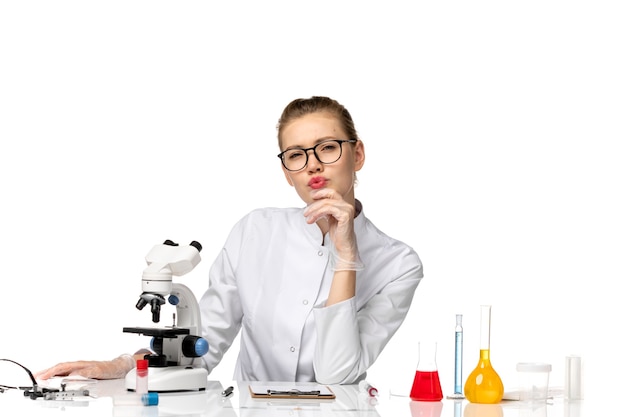 Vue de face femme médecin en costume médical blanc assis en face de la table avec des solutions de réflexion sur un espace blanc