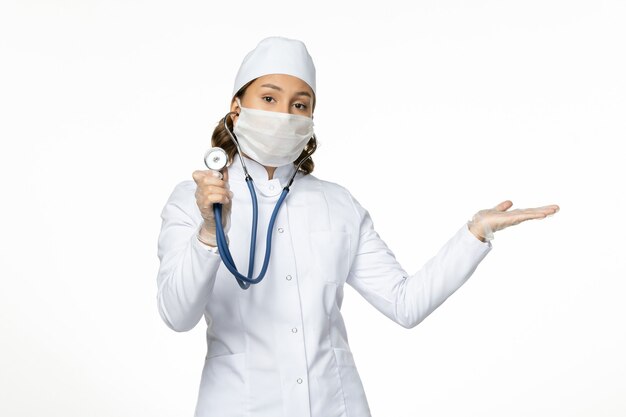 Vue de face femme médecin en costume médical blanc et masque à l'aide d'un stéthoscope sur fond blanc médecine de la maladie à virus pandémique