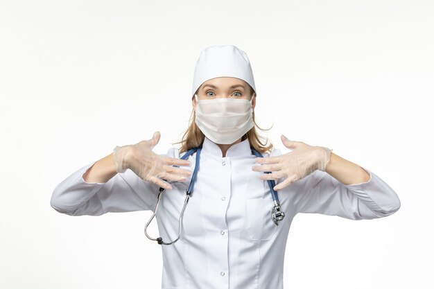 Vue de face femme médecin en costume médical blanc avec masque de protection en raison du coronavirus sur la maladie du mur blanc clair covid- maladie pandémique