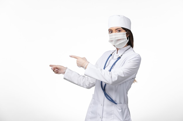 Vue de face femme médecin en costume médical blanc avec un masque en raison d'un coronavirus sur le mur blanc virus de la médecine pandémique covid-