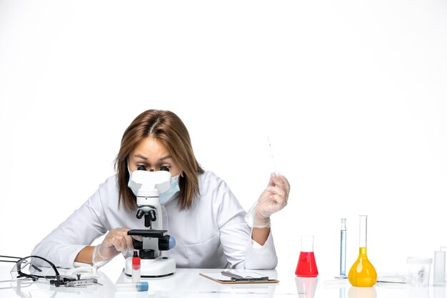 Vue de face femme médecin en costume médical blanc et masque en raison de covid à l'aide d'un microscope sur un espace blanc