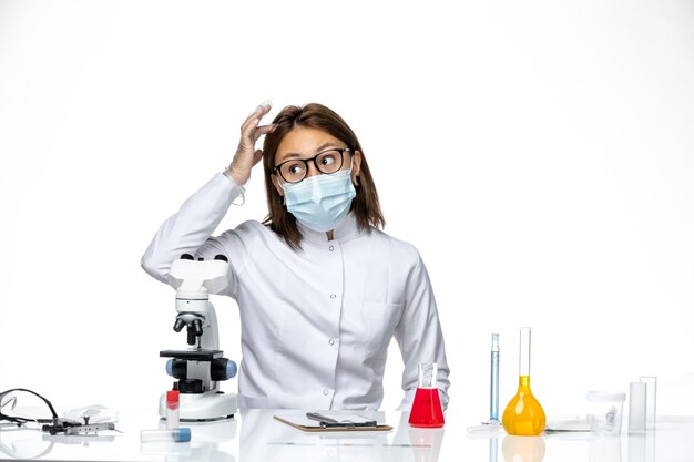 Vue de face femme médecin en costume médical blanc avec masque en raison de covid assis sur un espace blanc