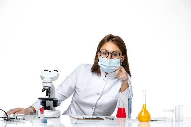 Vue de face femme médecin en costume médical blanc avec masque en raison de covid assis avec des solutions sur l'espace blanc clair