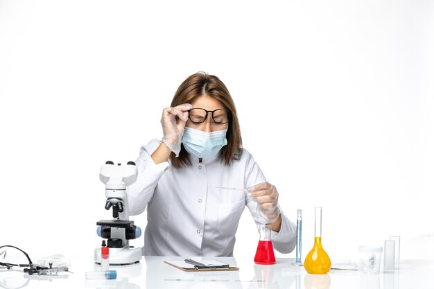 Vue de face femme médecin en costume médical blanc et masque en raison de covid travaillant avec des solutions sur un bureau blanc