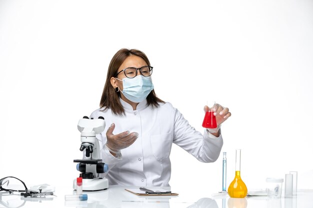 Vue de face femme médecin en costume médical blanc avec masque en raison de covid travaillant avec des solutions sur un bureau blanc