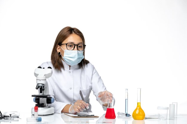 Vue de face femme médecin en costume médical blanc avec masque en raison de covid travaillant avec des solutions sur l'espace blanc