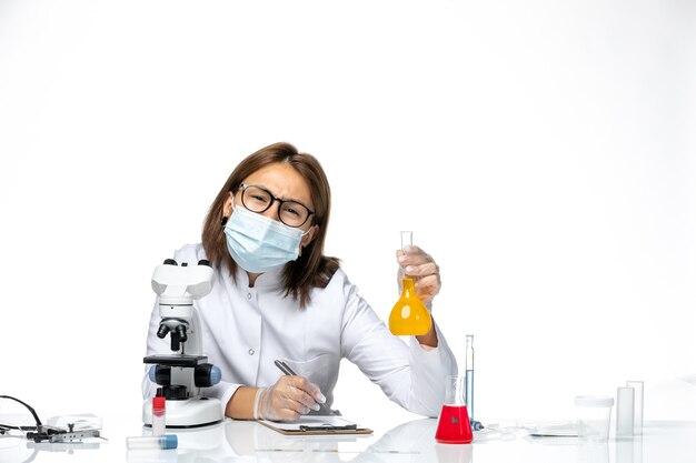 Vue de face femme médecin en costume médical blanc avec masque en raison de covid travaillant avec des solutions sur l'espace blanc