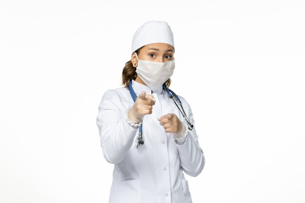 Vue de face femme médecin en costume médical blanc et avec masque en raison du coronavirus sur la maladie du virus du mur blanc clair pandémique covid