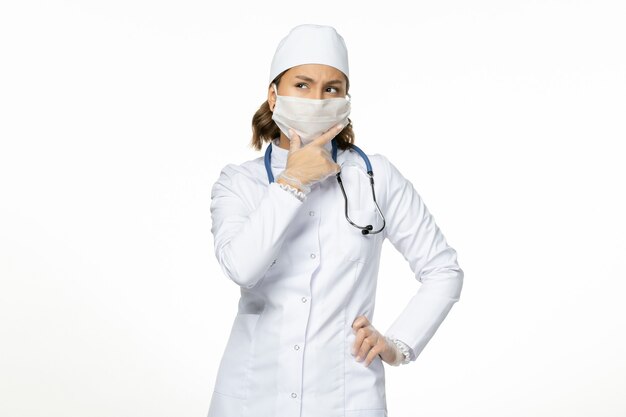 Vue de face femme médecin en costume médical blanc et avec masque en raison de la pensée du coronavirus sur la maladie à virus pandémique mur blanc covid