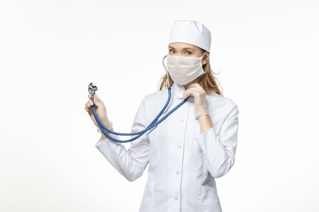 Vue de face femme médecin en costume médical blanc avec masque stérile en raison d'un coronavirus à l'aide d'un stéthoscope sur un bureau blanc maladie pandémique maladie covid-