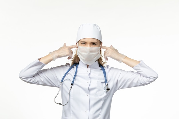 Vue de face femme médecin en costume médical blanc avec masque stérile en raison du coronavirus fermant ses oreilles sur le mur blanc maladie covid- maladie pandémique
