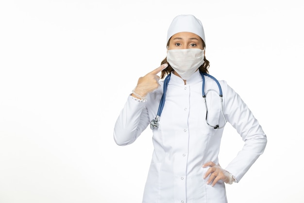 Vue de face femme médecin en costume médical blanc portant un masque en raison d'un coronavirus sur l'isolement de la maladie pandémique de mur blanc