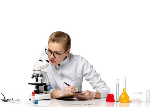 Vue de face femme médecin en costume médical blanc travaillant avec microscope sur espace blanc