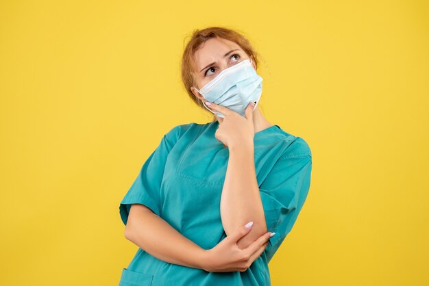 Vue de face de la femme médecin en costume médical et masque pensant sur mur jaune