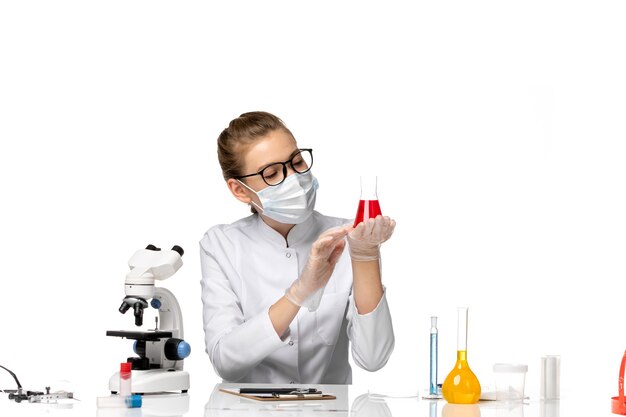 Vue de face femme médecin en costume médical avec masque en raison de covid travaillant avec des solutions sur un bureau blanc