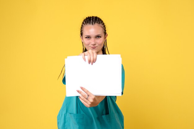 Vue de face de la femme médecin en costume médical avec des papiers sur mur jaune