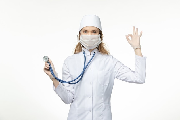 Vue de face femme médecin en costume médical portant un masque en raison de coronavirus tenant un stéthoscope sur le bureau blanc virus pandémique covid- maladie