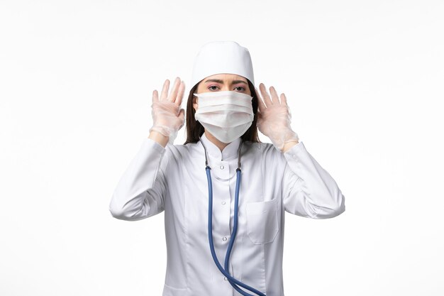 Vue de face femme médecin en costume médical stérile blanc avec masque en raison d'un coronavirus essayant d'entendre sur le mur blanc maladie covid- maladie à virus pandémique