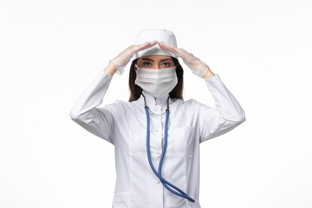 Vue de face femme médecin en costume médical stérile blanc avec masque en raison de coronavirus posant sur mur blanc maladie covid- maladie à virus pandémique