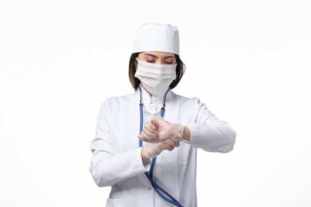 Vue de face femme médecin en costume médical stérile blanc avec masque en raison de coronavirus regardant son poignet sur mur blanc maladie maladie virus pandémique covid-