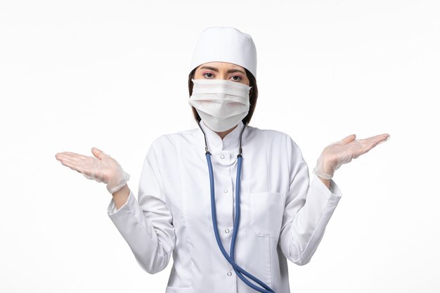 Vue de face femme médecin en costume médical stérile blanc avec masque en raison du coronavirus sur le virus pandémique de la maladie du mur blanc covid-