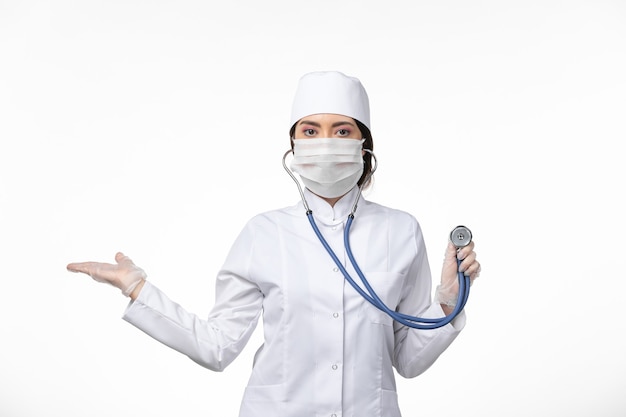 Vue de face femme médecin en costume médical stérile blanc et portant un masque en raison de covid- à l'aide d'un stéthoscope sur le sol blanc virus de la maladie covid- maladie pandémique