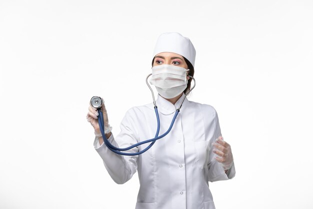 Vue de face femme médecin en costume médical stérile blanc et portant un masque en raison de covid- à l'aide d'un stéthoscope sur le virus de la maladie du mur blanc clair covid- maladie pandémique