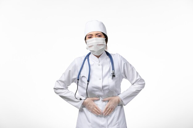 Vue de face femme médecin en costume médical stérile blanc et portant un masque en raison de covid- ayant mal au ventre sur le virus de la maladie du mur blanc covid- maladie pandémique