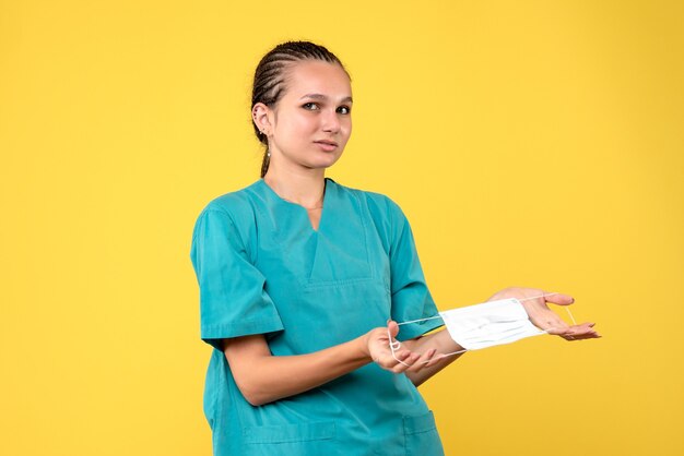 Vue de face de la femme médecin en costume médical tenant un masque stérile sur mur jaune