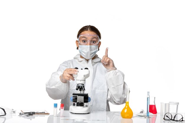 Vue de face femme médecin en costume spécial et portant un masque travaillant au microscope sur le coronavirus pandémique du virus covid de plancher blanc