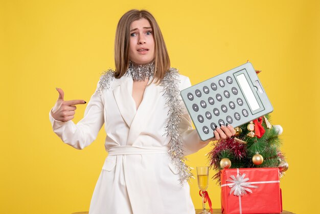 Vue de face femme médecin debout et tenant la calculatrice sur fond jaune avec arbre de Noël et coffrets cadeaux