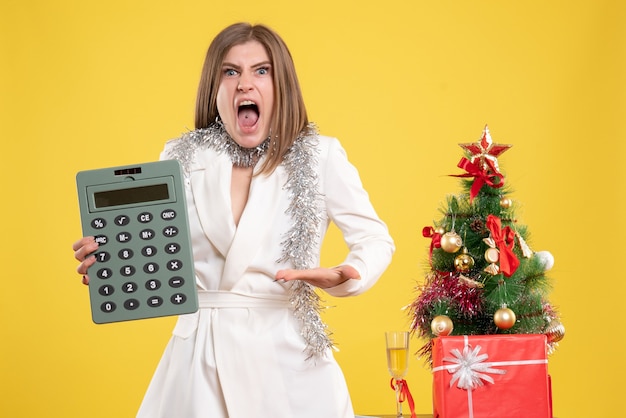 Vue de face femme médecin debout et tenant en colère calculatrice sur fond jaune avec arbre de Noël et coffrets cadeaux