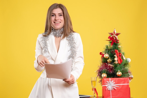 Vue De Face Femme Médecin Debout Et Tenant Des Documents Sur Fond Jaune Avec Arbre De Noël Et Coffrets Cadeaux