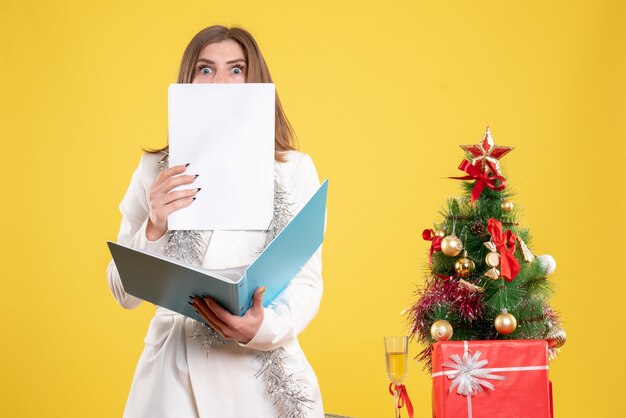 Vue de face femme médecin debout et tenant des documents sur fond jaune avec arbre de Noël et coffrets cadeaux