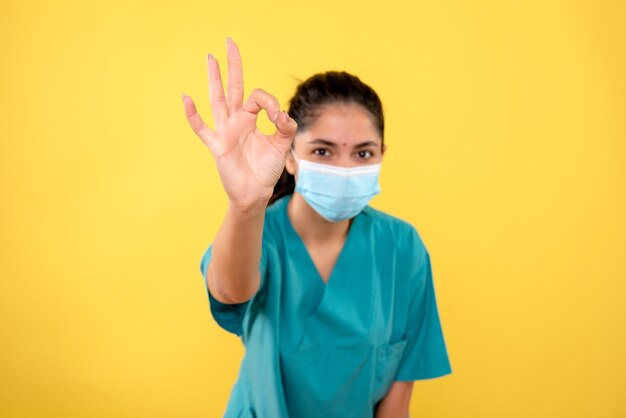 Vue de face de la femme médecin avec masque faisant signe okey devant l'avant sur le mur jaune