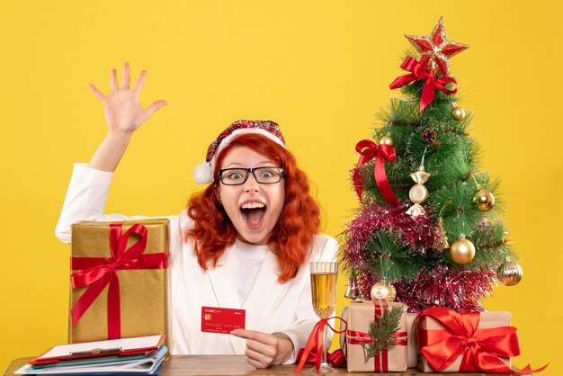 Vue de face femme médecin tenant une carte bancaire autour de cadeaux de Noël et arbre