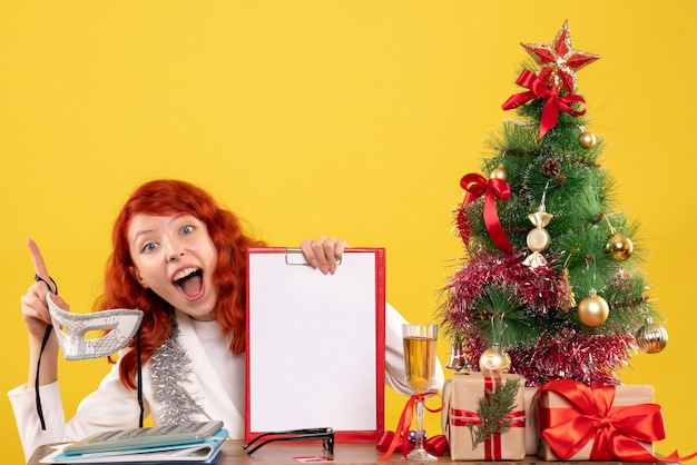 Vue de face femme médecin tenant un masque et une note de fichier autour de l'arbre de Noël