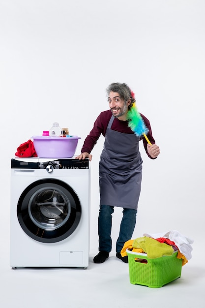 Photo gratuite vue de face femme de ménage exaltée tenant un plumeau debout près du panier à linge de la machine à laver sur fond blanc