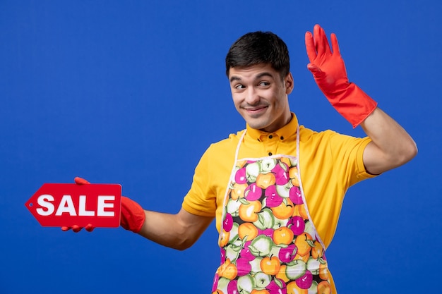 Photo gratuite vue de face d'une femme de ménage souriante en t-shirt jaune et tablier tenant une pancarte de vente sur le mur bleu