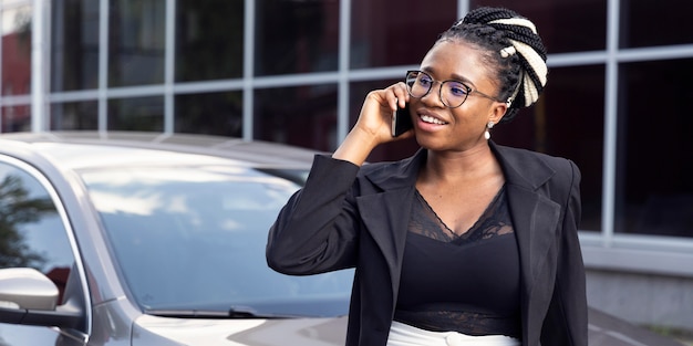 Vue de face de femme parlant sur smartphone tout en s'appuyant contre sa voiture