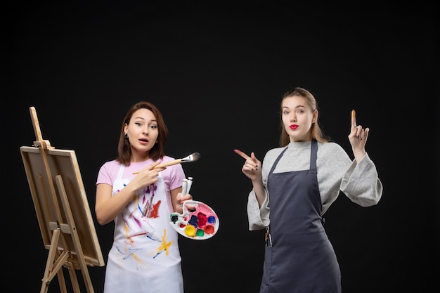 Vue de face femme peintre dessin sur chevalet avec d'autres femmes sur mur noir photo couleur art photos artiste travail de peinture