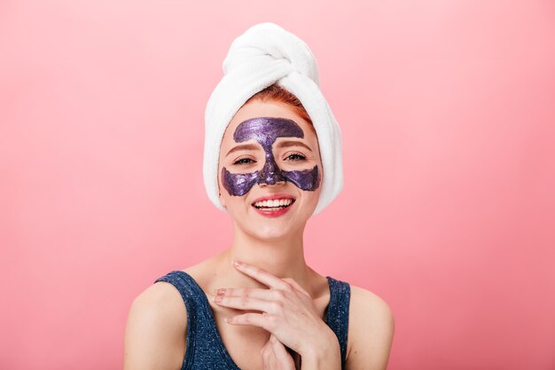 Vue de face de la femme qui rit dans une serviette et un masque isolé sur fond rose. Fille heureuse faisant le traitement du visage avec le sourire.