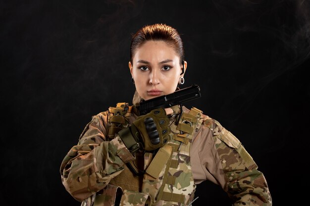 Vue de face d'une femme soldat avec une arme à feu en uniforme sur un mur noir